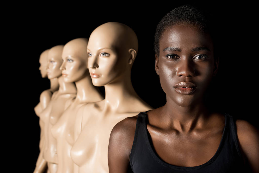serious african american girl looking at camera while standing with dummies on black - Photo, Image
