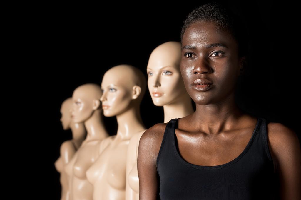 serious african american woman in swimsuit standing in row with mannequins on black - Photo, Image