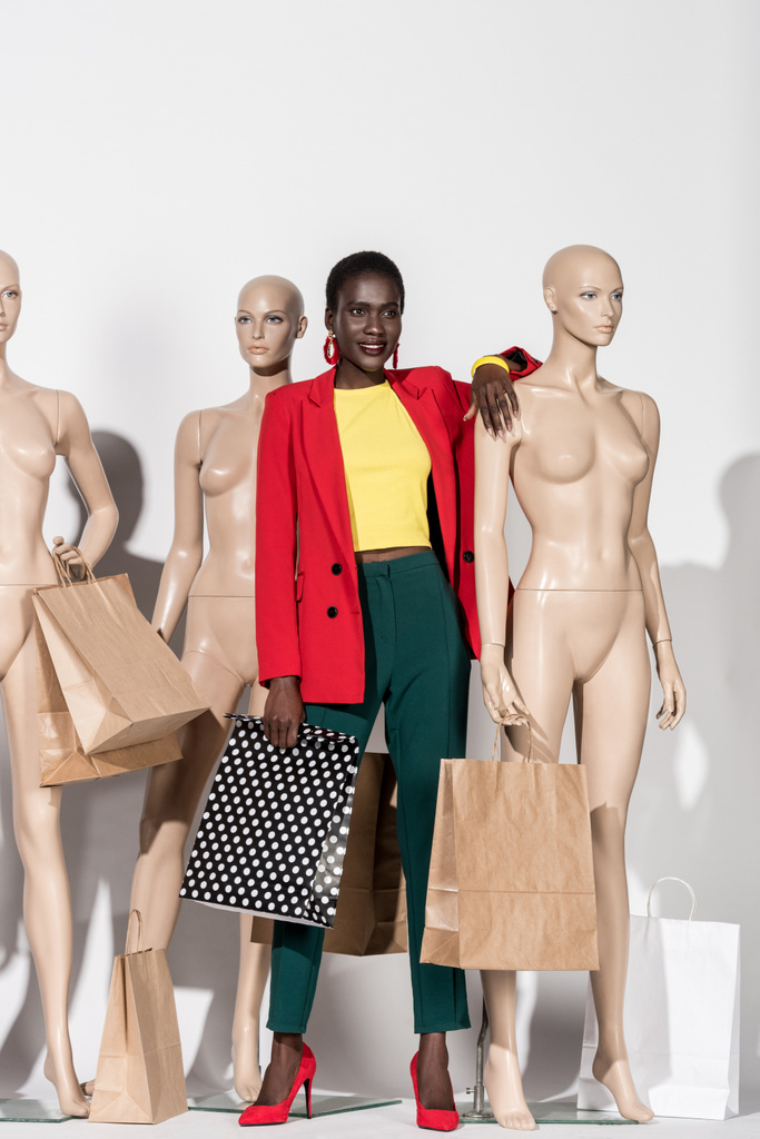 visão comprimento total da bela menina americana africana sorridente de pé entre manequins com sacos de compras em branco
  - Foto, Imagem