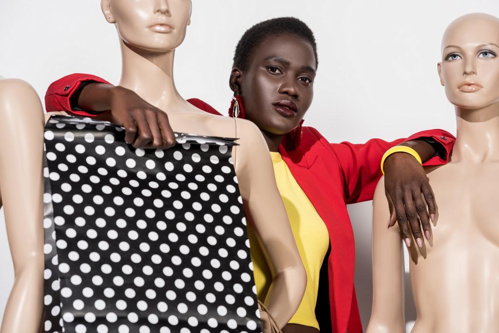 mladá módní africká americká žena drží nákupní tašku a při pohledu na kameru naklánět na figuríny na bílém  - Fotografie, Obrázek