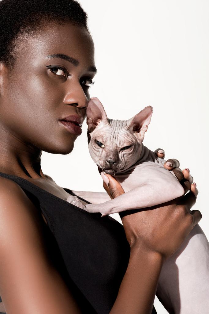 vista de cerca de la hermosa mujer afroamericana en traje de baño sosteniendo gato esfinge y mirando a la cámara aislada en blanco
 - Foto, Imagen