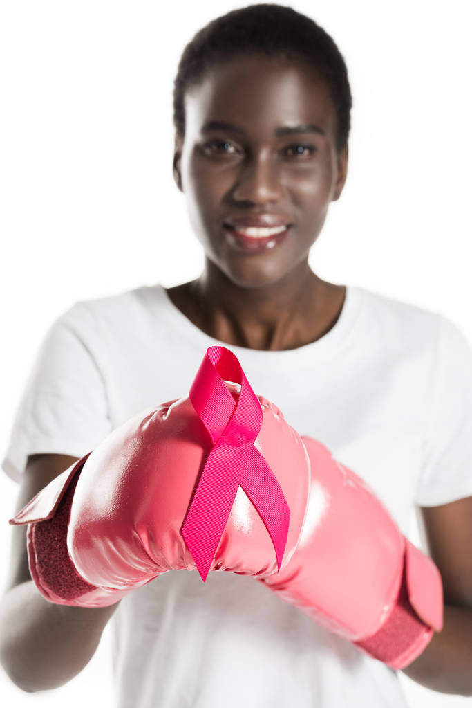 close-up view of young african american woman in boxing gloves holding pink ribbon and smiling at camera isolated on white, breast cancer concept - Photo, Image