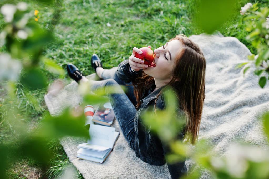 春に対する格子縞の上に座ってジーンズで若いブルネットの少女の木の花し本を読み、リンゴを食べる. - 写真・画像