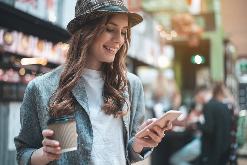 Улыбающаяся девушка с кофе использует смартфон
 - Фото, изображение
