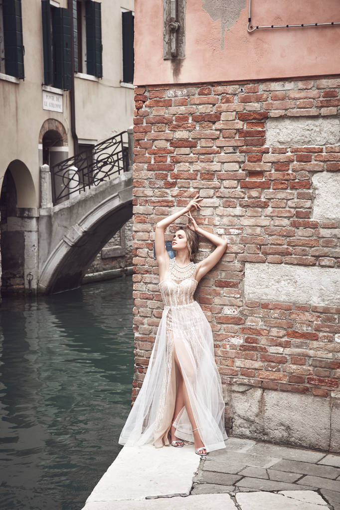 Belle mariée avec maquillage nu, vêtue d'une élégante robe de luxe en ivoire et d'une coiffure désordonnée, posant près du canal de Venise
 - Photo, image