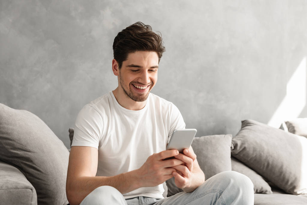 Beau homme en t-shirt basique souriant et tenant le téléphone portable dans les mains tout en étant assis sur le canapé dans le salon - Photo, image