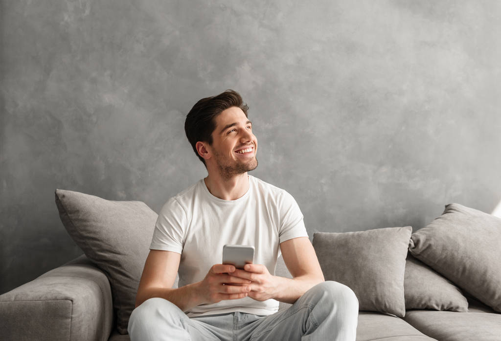 Hombre sonriente contento en camiseta básica sosteniendo el teléfono inteligente en las manos y mirando a un lado con mirada feliz mientras está sentado en el sofá dentro
 - Foto, imagen