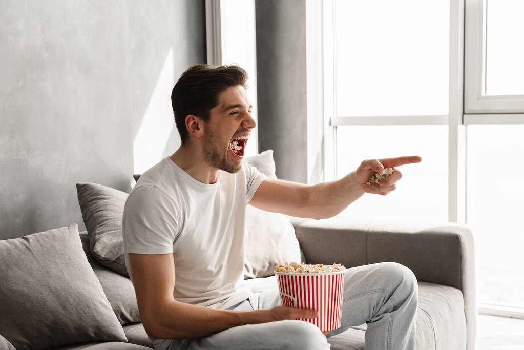 ポップコーンを食べながらリビング ルームとテレビでジェスチャー指でソファーに座っていた恍惚とした幸せな男 - 写真・画像