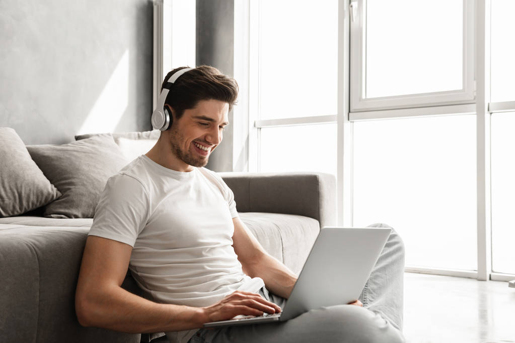 bärtiger Mann um die 30 in einfacher Kleidung sitzt zu Hause auf dem Boden und hört Musik am Laptop mit drahtlosen Kopfhörern - Foto, Bild