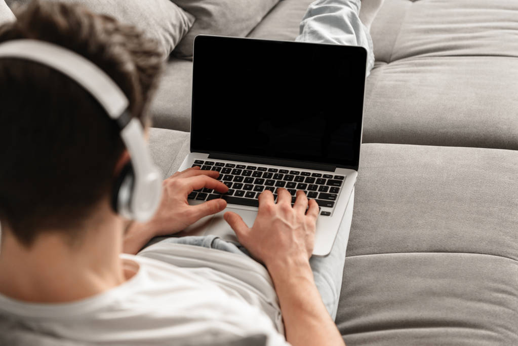 Фото ззаду юнака 30s в повсякденному одязі, що друкується на ноутбуці під час роботи вдома з навушниками
 - Фото, зображення