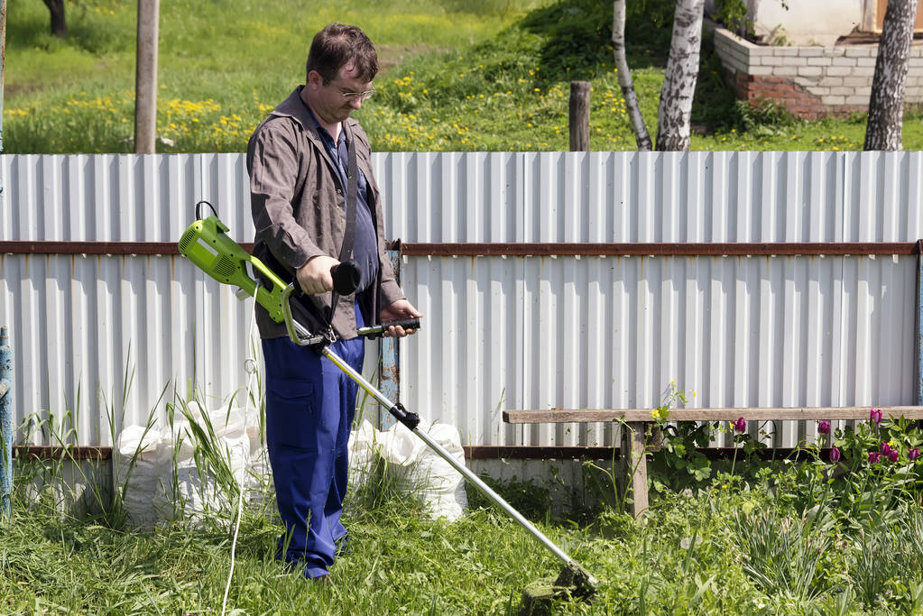 un uomo in bicchieri con un trimer falcia l'erba nel cortile di una casa nel villaggio, porta ordine nel territorio del paese
 - Foto, immagini