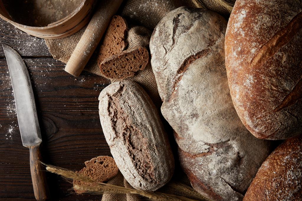 vista elevada de vários tipos de pão, trigo, rolo de pino, peneira e saco na mesa de madeira
  - Foto, Imagem