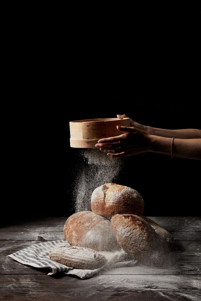 荒布を着た黒の背景に分離されたパンのさまざまな種類の小麦粉をふるう女性のベイカーのクロップ撮影  - 写真・画像