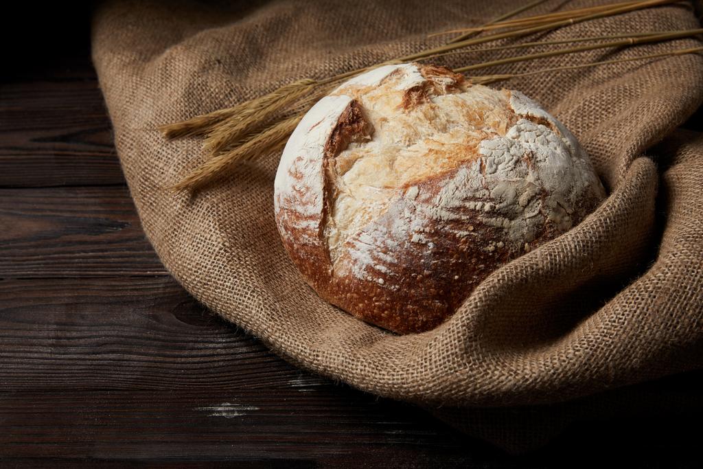 Nahaufnahme von Brot, Weizen und Sacktuch auf rustikalem Holztisch  - Foto, Bild