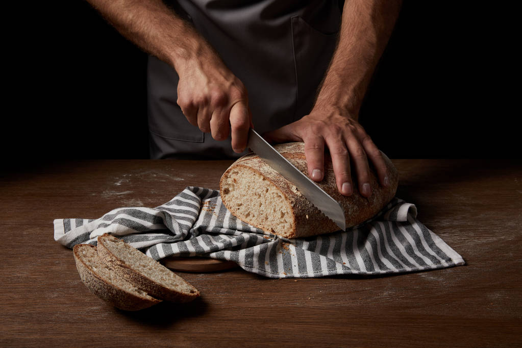Schnappschuss eines Bäckers, der Brot mit einem Messer auf einem Sacktuch auf Holztisch schneidet - Foto, Bild