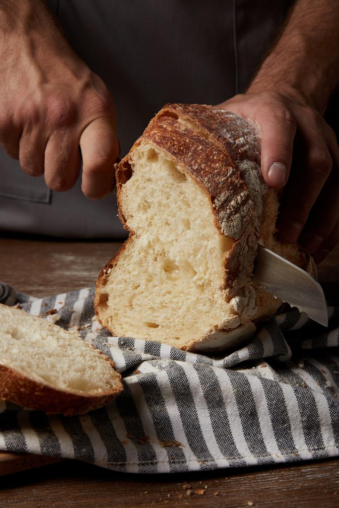 Colpo ritagliato di panettiere maschio che taglia il pane con un coltello sul sacco sul tavolo di legno
 - Foto, immagini