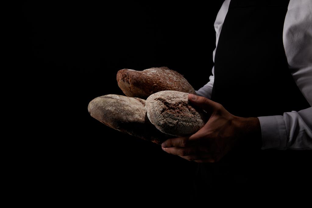 abgeschnittenes Bild eines männlichen Bäckers in Schürze mit verschiedenen Brotsorten auf schwarzem Hintergrund  - Foto, Bild