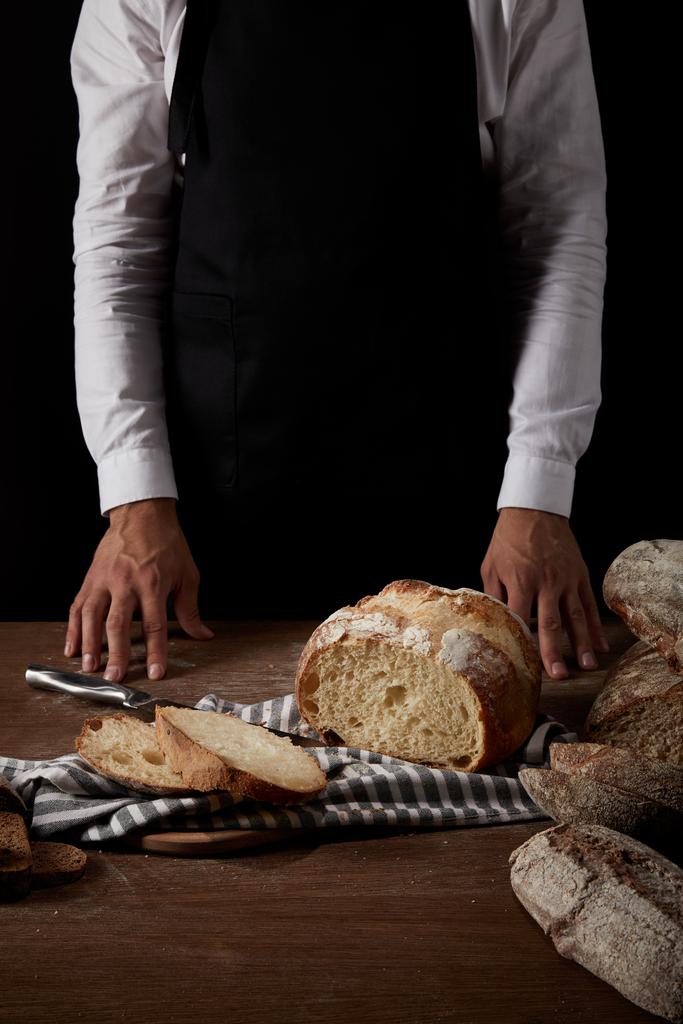 imagem cortada de padeiro macho em avental de pé perto da mesa com pão, faca e saco
 - Foto, Imagem