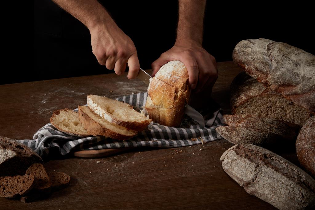 schot van mannelijke baker snijden brood bijgesneden door mes op zak op houten tafel - Foto, afbeelding
