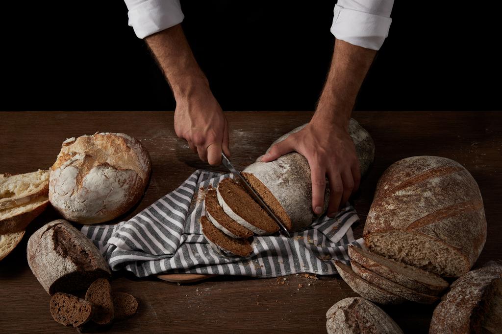 cropped image of male baker cutting bread by knife on sackcloth on wooden table - Photo, Image