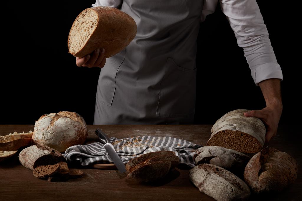częściowy widok mężczyzna baker w fartuch, trzymając chleb na stole z noża, chleb i worze  - Zdjęcie, obraz