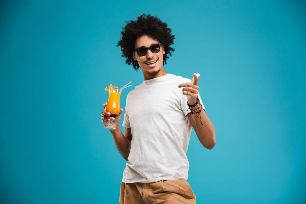 Foto van de knappe jonge Afrikaanse krullend man die boven de blauwe achtergrond houden cocktail wijzen geïsoleerd staan. - Foto, afbeelding
