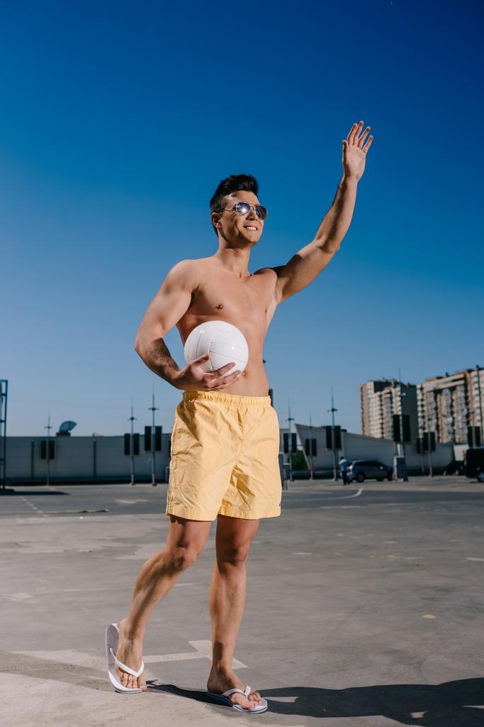 glücklicher Mann ohne Hemd hält Volleyball und winkt mit der Hand auf dem Parkplatz - Foto, Bild