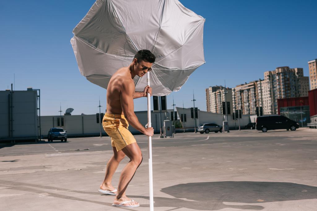 masculine young shirtless man putting beach umbrella in asphalt on parking - Photo, Image