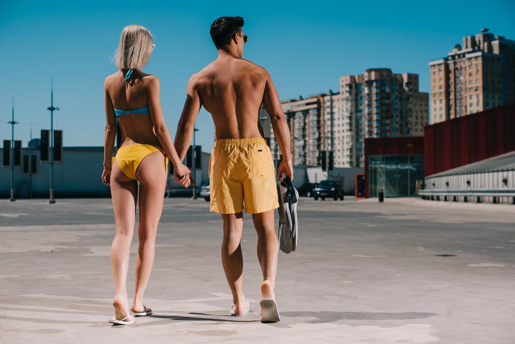 rear view of young couple in bikini and swimming shorts with flippers walking on parking - Photo, Image