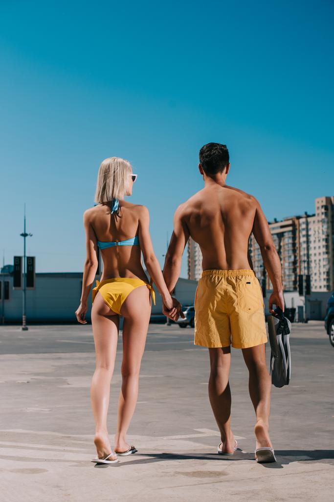 vista trasera de pareja joven atractiva en bikini y pantalones cortos de natación con aletas caminando en el estacionamiento
 - Foto, imagen