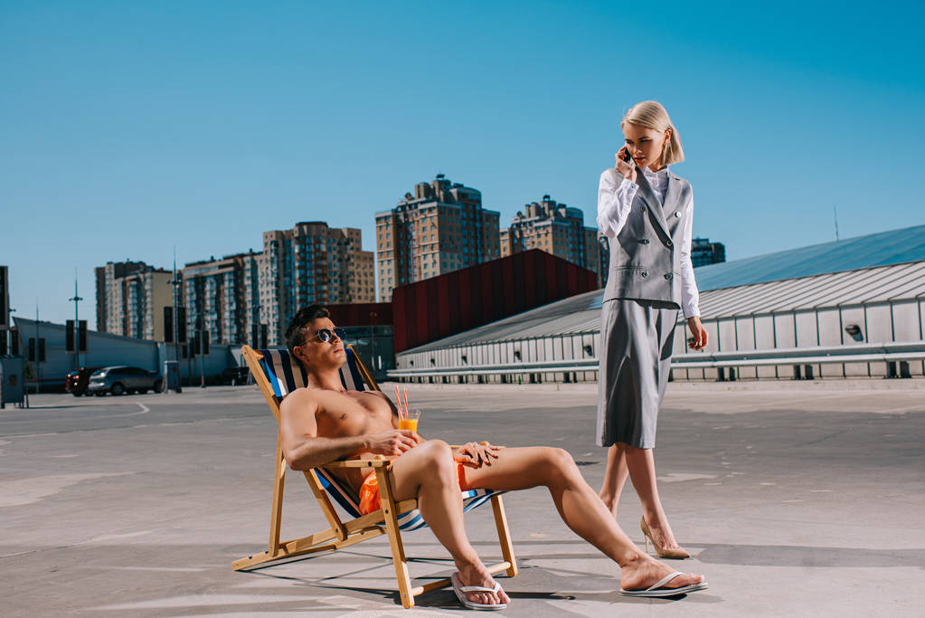 komea nuori mies rentoutuu aurinkotuolissa, kun hänen naispuolinen pomonsa puvussa hänen vieressään ja puhuu puhelimitse pysäköinnissä
 - Valokuva, kuva