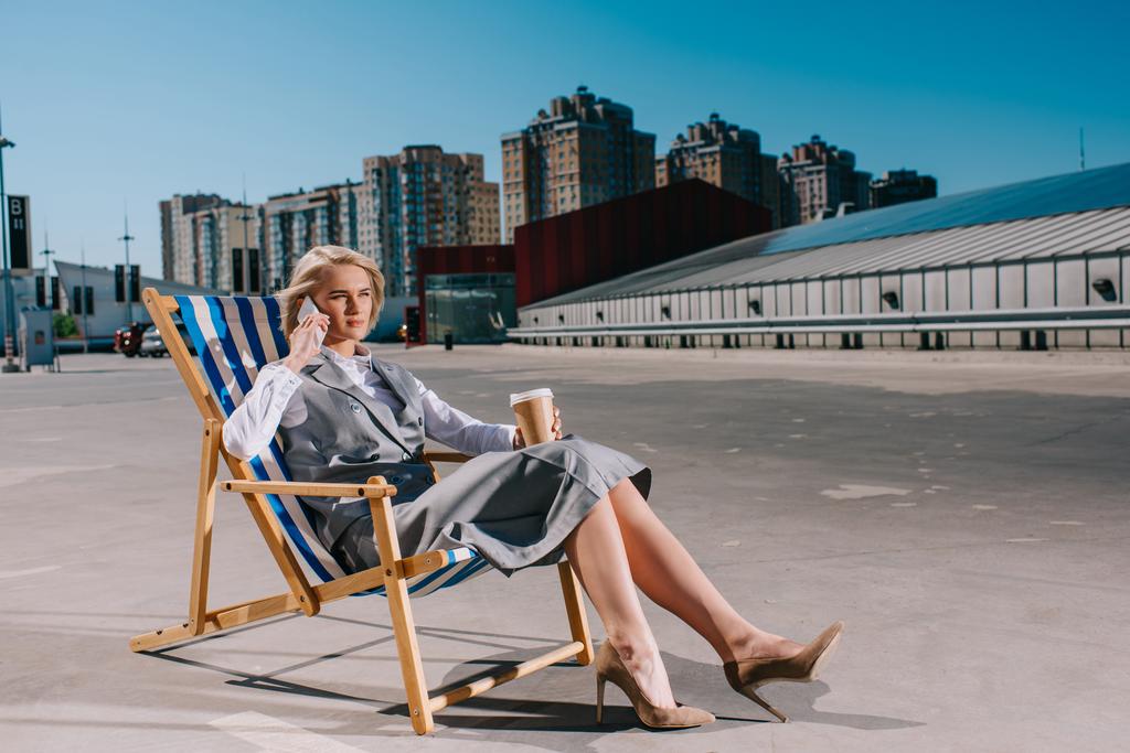 attraktive junge Geschäftsfrau in formeller Kleidung sitzt in einer Sonnenliege mit Coffee to go und telefoniert auf dem Parkplatz - Foto, Bild