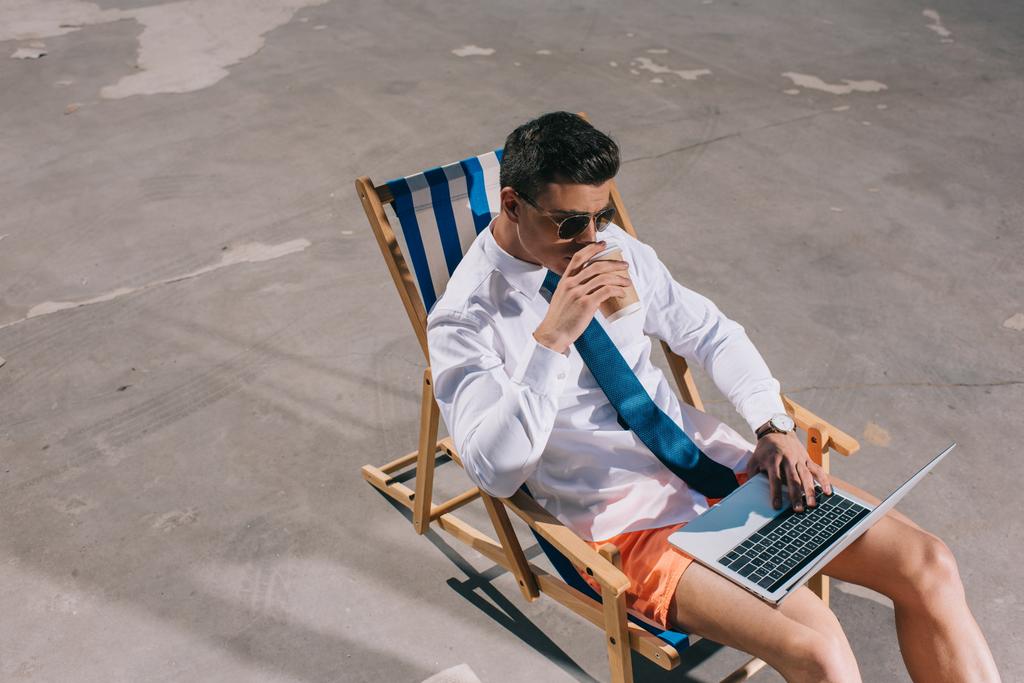 vista de ángulo alto de hombre de negocios joven en pantalones cortos que trabajan con el ordenador portátil mientras está sentado en la tumbona sobre asfalto y beber café para ir
 - Foto, imagen