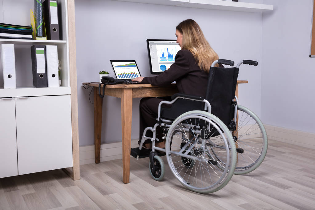 Taustakuva vammaisesta liikenaisesta istumassa pyörätuolilla työskentelemässä tietokoneella
 - Valokuva, kuva