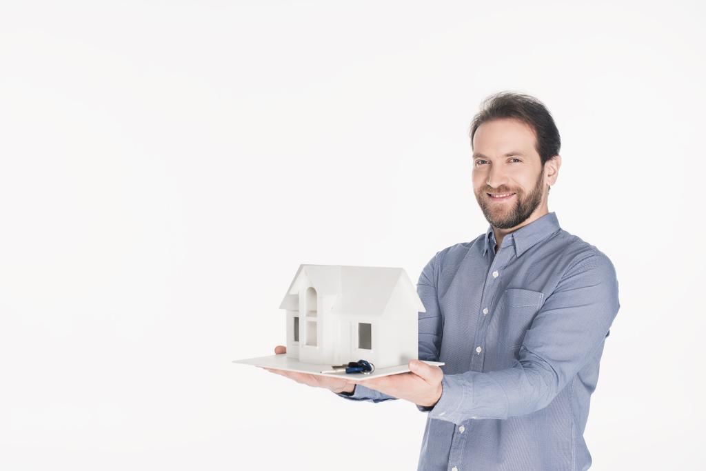 πορτρέτο του χαμογελώντας γενειοφόρο άτομο που κρατά το σπίτι μοντέλο και τα κλειδιά στα χέρια που απομονώνονται σε λευκό - Φωτογραφία, εικόνα