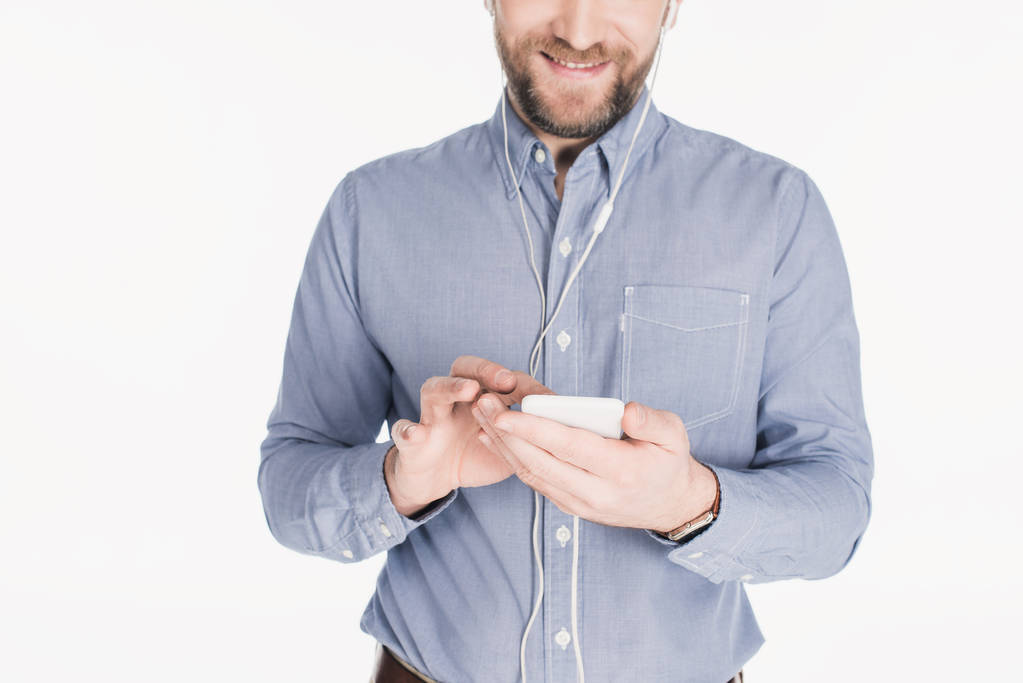 částečný pohled usmívající se vousatý muž v sluchátka pomocí chytrého izolované na bílém - Fotografie, Obrázek