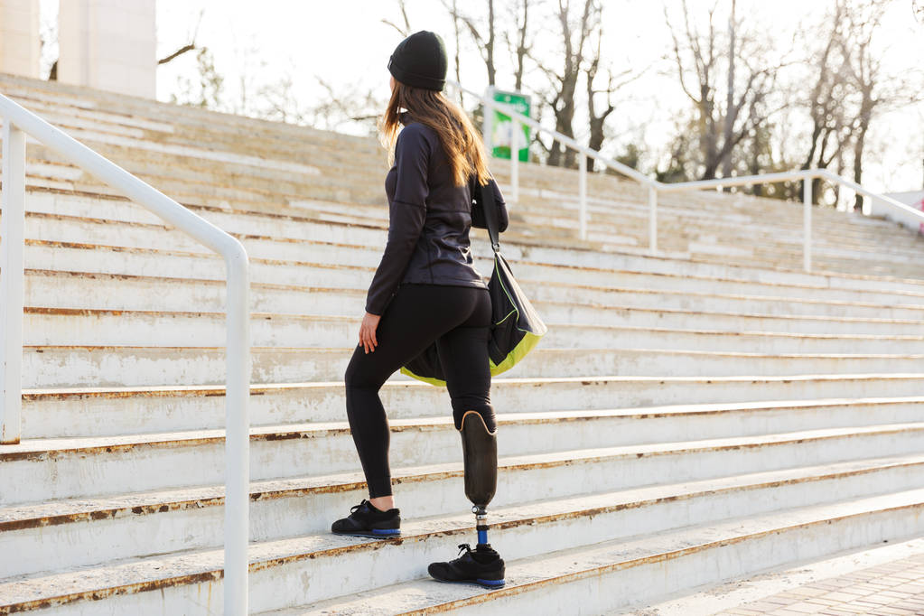 Foto de la espalda de una joven atlética discapacitada con una pierna protésica en ropa deportiva subiendo las escaleras al aire libre.
 - Foto, Imagen