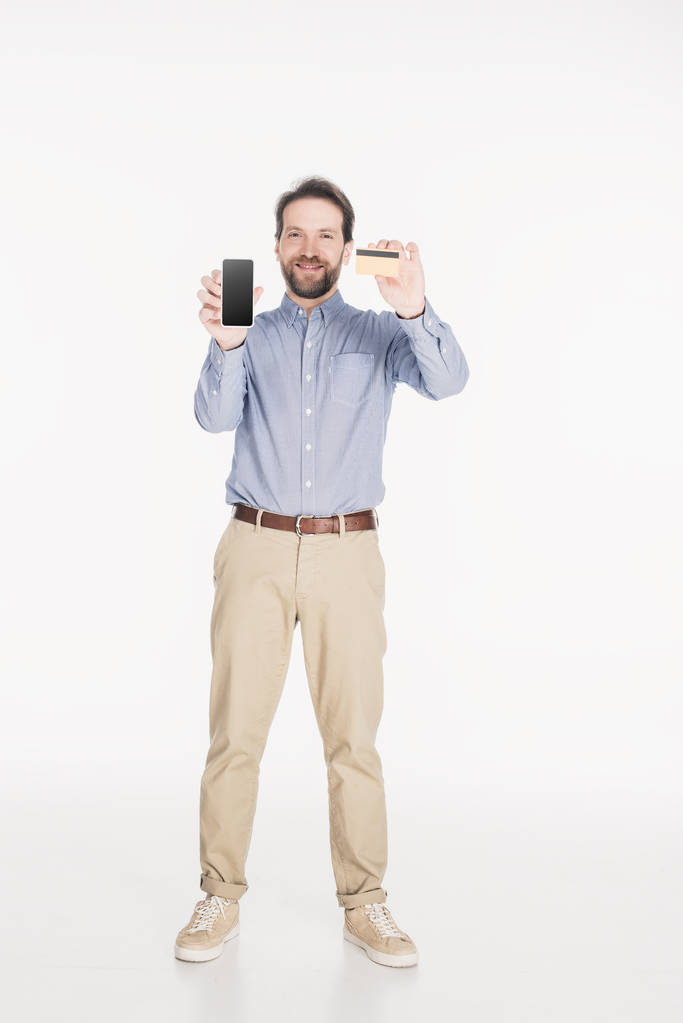 Lächelnder bärtiger Mann zeigt Smartphone mit leerem Bildschirm und Kreditkarte in weiß isolierten Händen - Foto, Bild