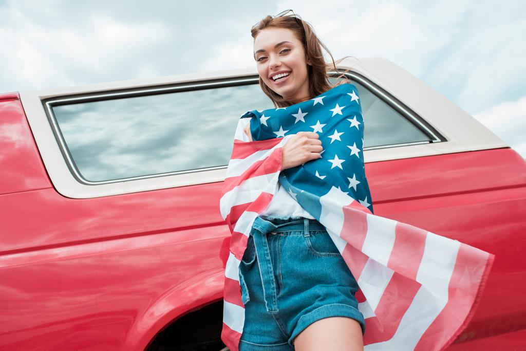 χαρούμενο κορίτσι τυλιγμένο σε αμερικανική σημαία στέκεται κοντά σε κόκκινο αυτοκίνητο - Φωτογραφία, εικόνα