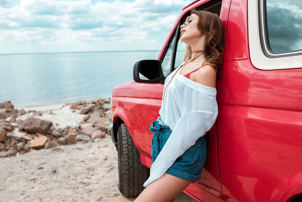 fröhliche junge Frau posiert mit rotem Auto während Fahrt am Meer - Foto, Bild