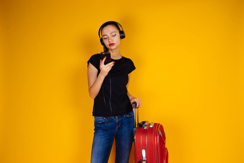 una joven esbelta en jeans escuchando música en los auriculares, esperando su avión con una maleta grande, se va de viaje
 - Foto, imagen