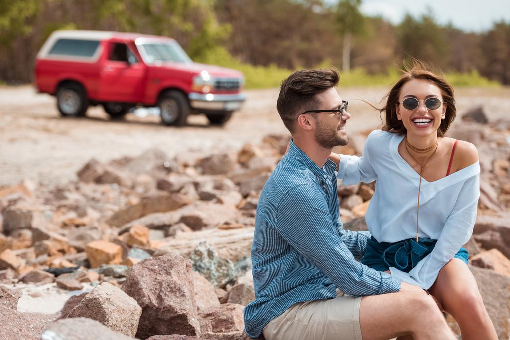 couple souriant assis sur des rochers avec jeep rouge sur le fond
 - Photo, image