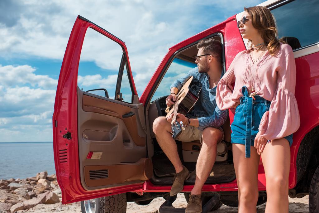 young man playing acoustic guitar near car with girlfriend on road trip - Photo, Image