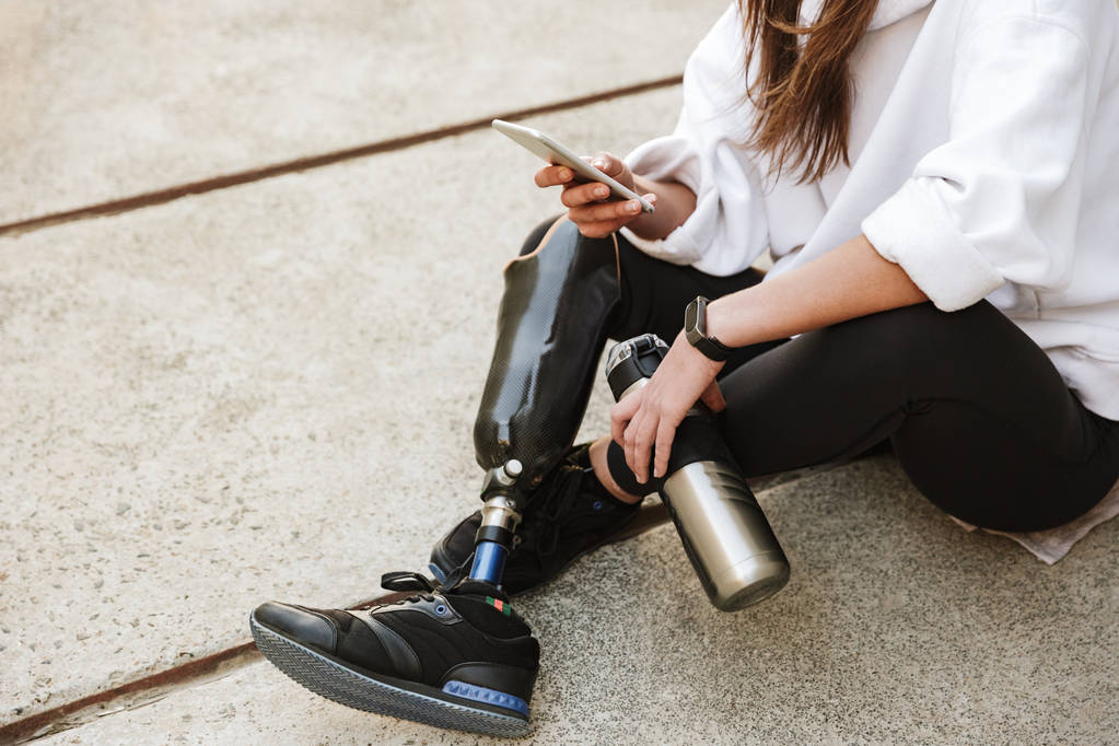 zugeschnittenes Foto einer behinderten Frau in Streetwear mit bionischem Bein, die im Freien auf Betonboden sitzt und ihr Handy benutzt - Foto, Bild