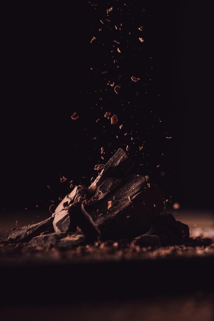 Nahaufnahme von geriebener dunkler Schokolade, die auf Schokoladenstücke auf schwarzem Hintergrund fällt  - Foto, Bild