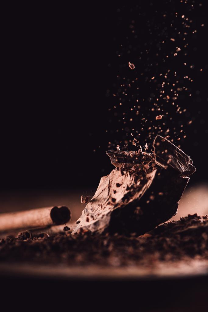 Close-up shot van kaneelstokje en geraspte chocolade vallen op stukjes chocolade op zwarte achtergrond  - Foto, afbeelding