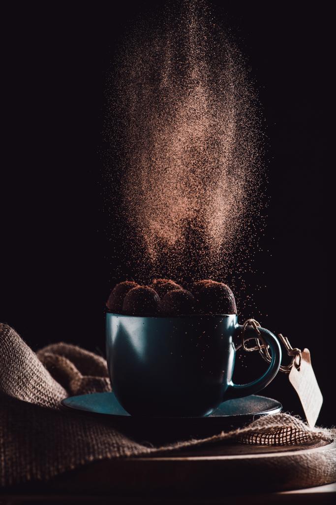 close up tiro de chocolate ralado caindo em trufas na xícara de café em pano de saco e placa de corte em fundo preto
  - Foto, Imagem