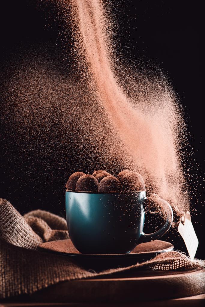 primer plano vista de chocolate rallado cayendo sobre trufas en taza de café sobre tela de saco y tabla de cortar sobre fondo negro
  - Foto, Imagen