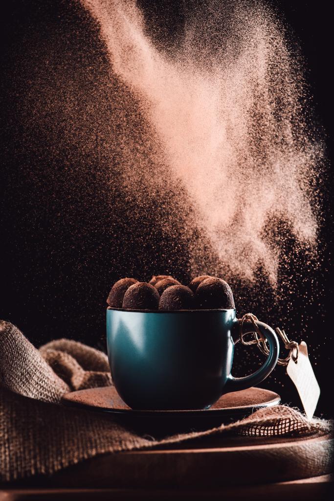 imagem de close-up de chocolate ralado caindo em trufas na xícara de café em pano de saco e placa de corte em fundo preto
  - Foto, Imagem