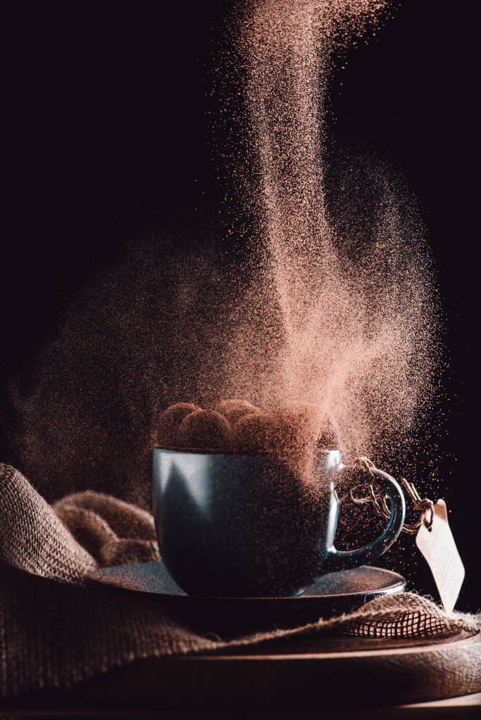  Nahaufnahme von geriebener Schokolade, die auf Trüffeln in einer Tasse Kaffee auf Sacktuch und Schneidebrett auf schwarzem Hintergrund fällt  - Foto, Bild